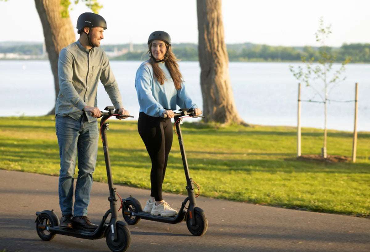 Dos personas con casco circulan en patinetes eléctricos NAVEE por un camino junto a un lago
