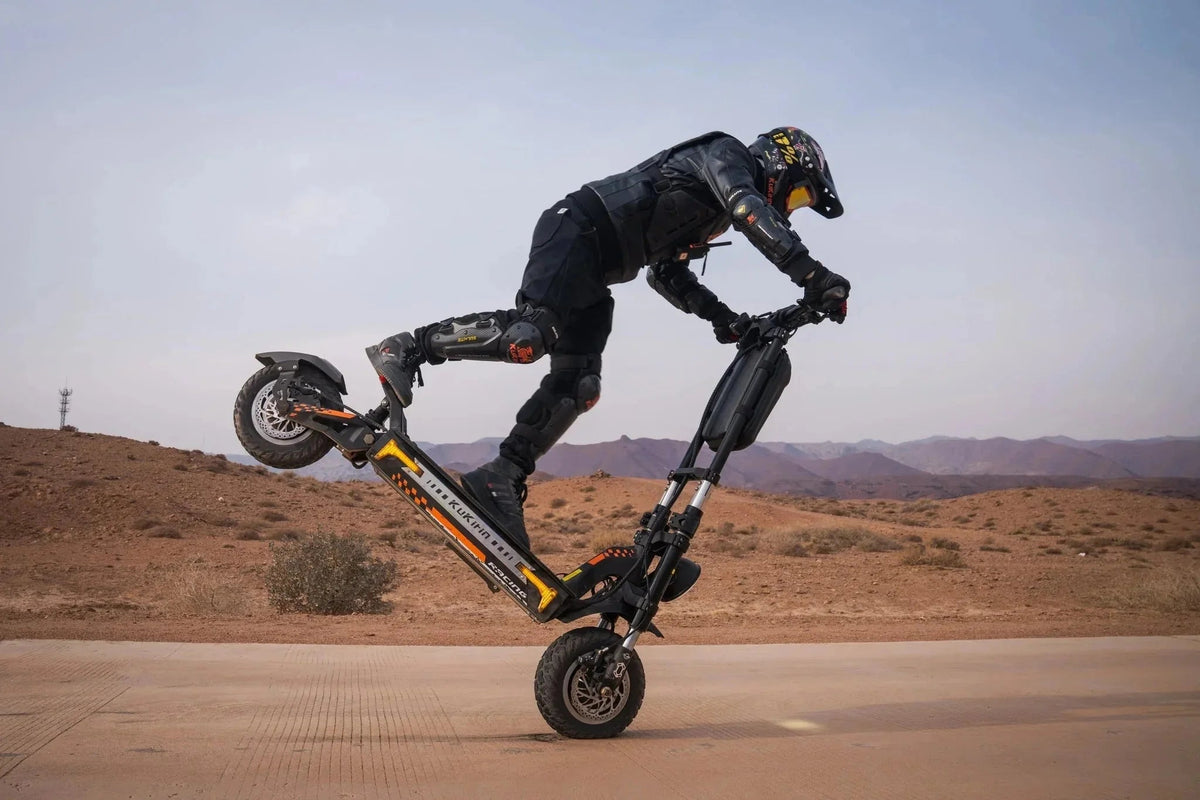 Piloto con casco y protecciones levanta la rueda trasera de un patinete eléctrico KuKirin negro y naranja en el desierto