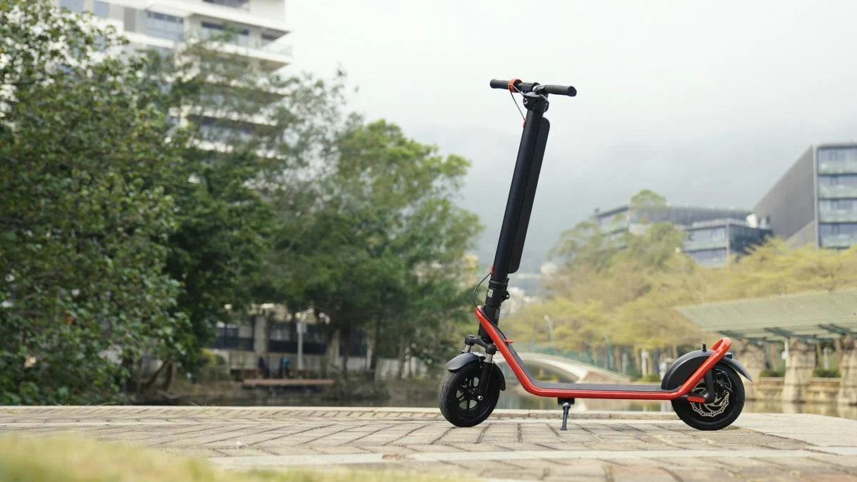 Patinete eléctrico HX negro con cuadro rojo sobre su pata de apoyo en un paseo adoquinado, con árboles y edificios detrás