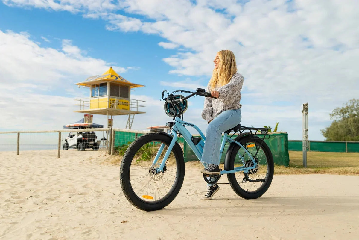 Mujer en bicicleta eléctrica parada junto a la playa, con el casco en el manillar – normativa de la bicicleta eléctrica