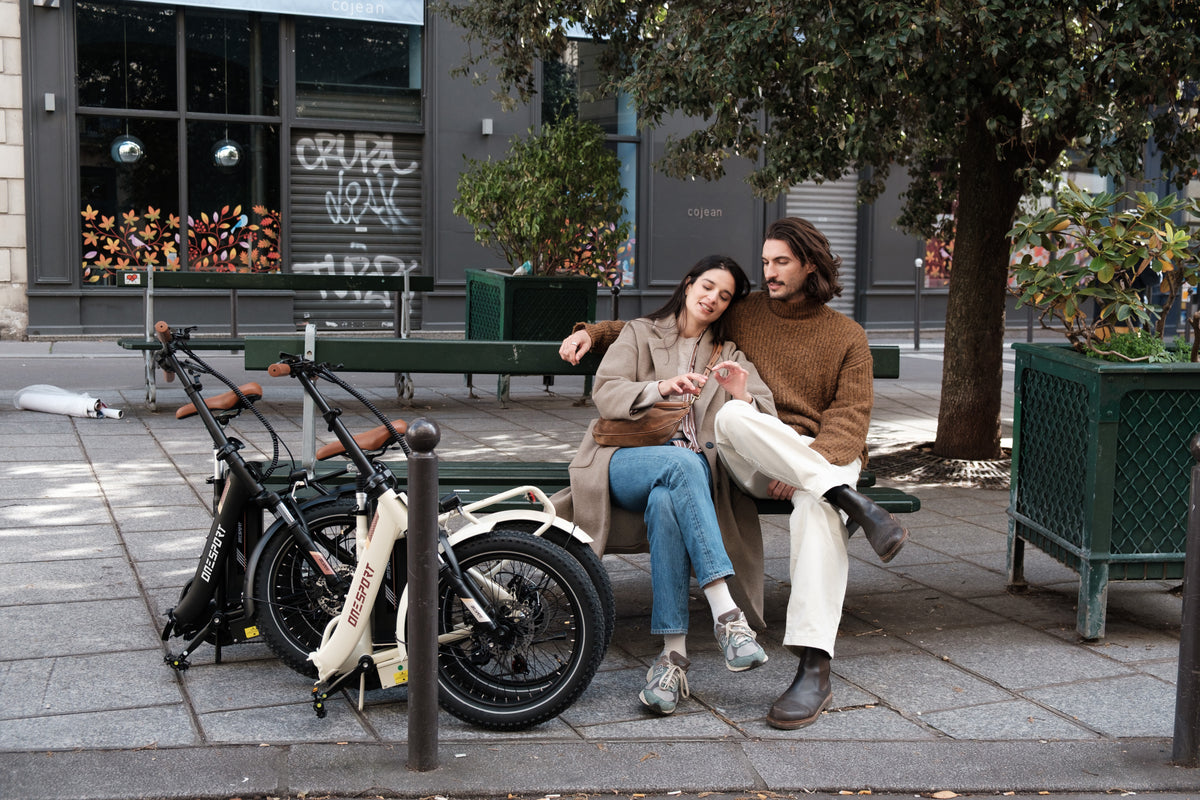 Una pareja sentada en un banco junto a dos bicicletas OneSport plegadas, una beige y una negra