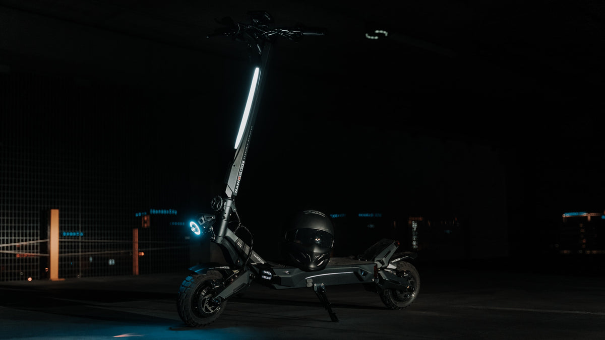 Patinete eléctrico negro aparcado sobre la pata de cabra en un garaje oscuro, con un casco sobre la plataforma