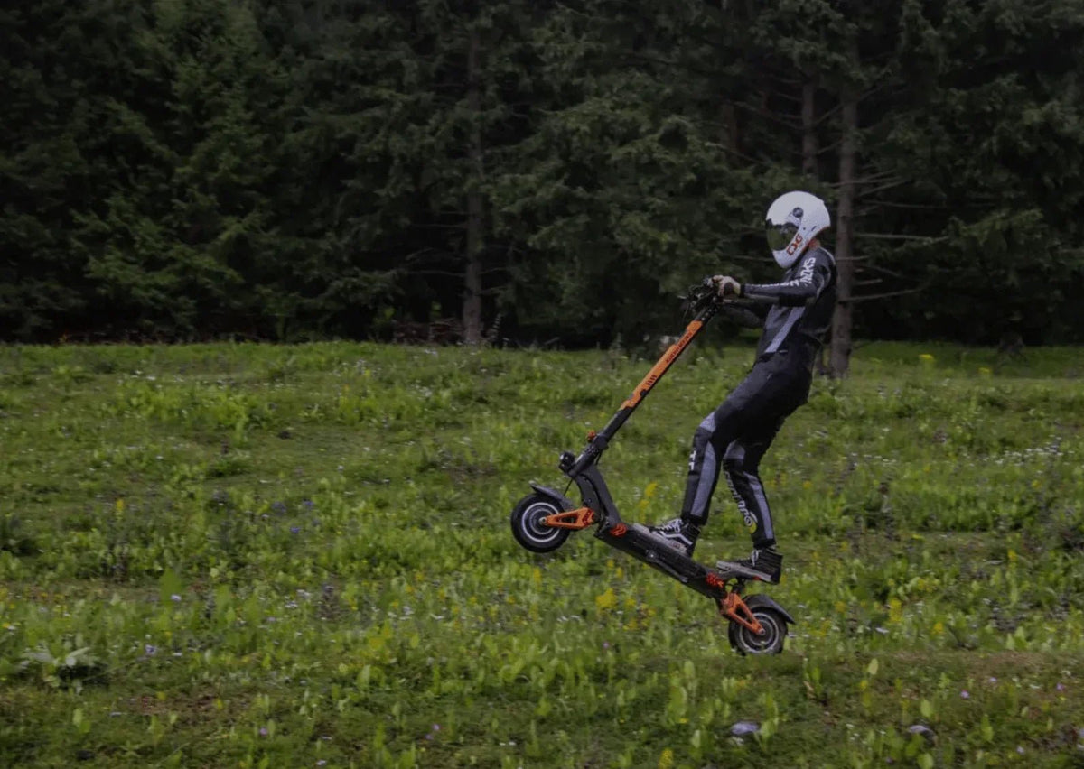 Piloto con casco blanco y mono salta con un patinete eléctrico negro y naranja sobre un prado junto a un bosque de abetos