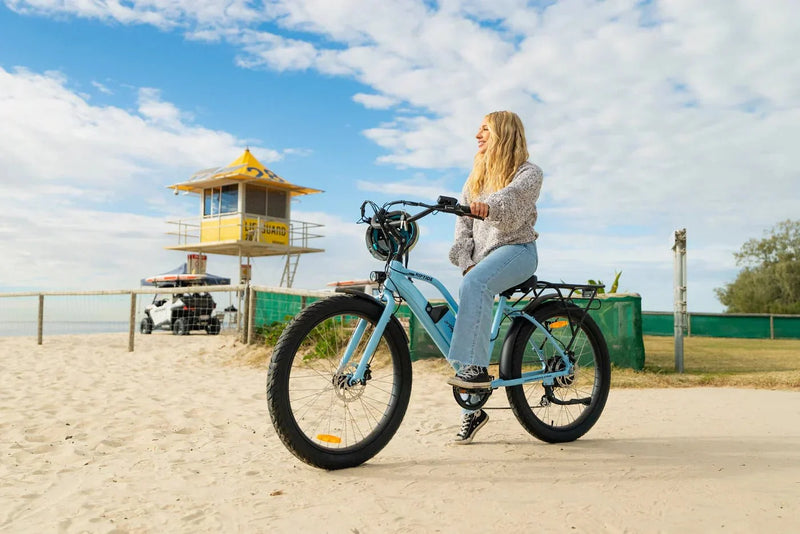 Mujer en bicicleta eléctrica parada junto a la playa, con el casco en el manillar – normativa de la bicicleta eléctrica