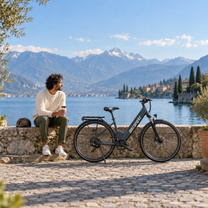 Bicicleta eléctrica OneSport OT12 Pro en un muelle empedrado junto a un lago, con un hombre sentado en el muro al lado y montañas al fondo