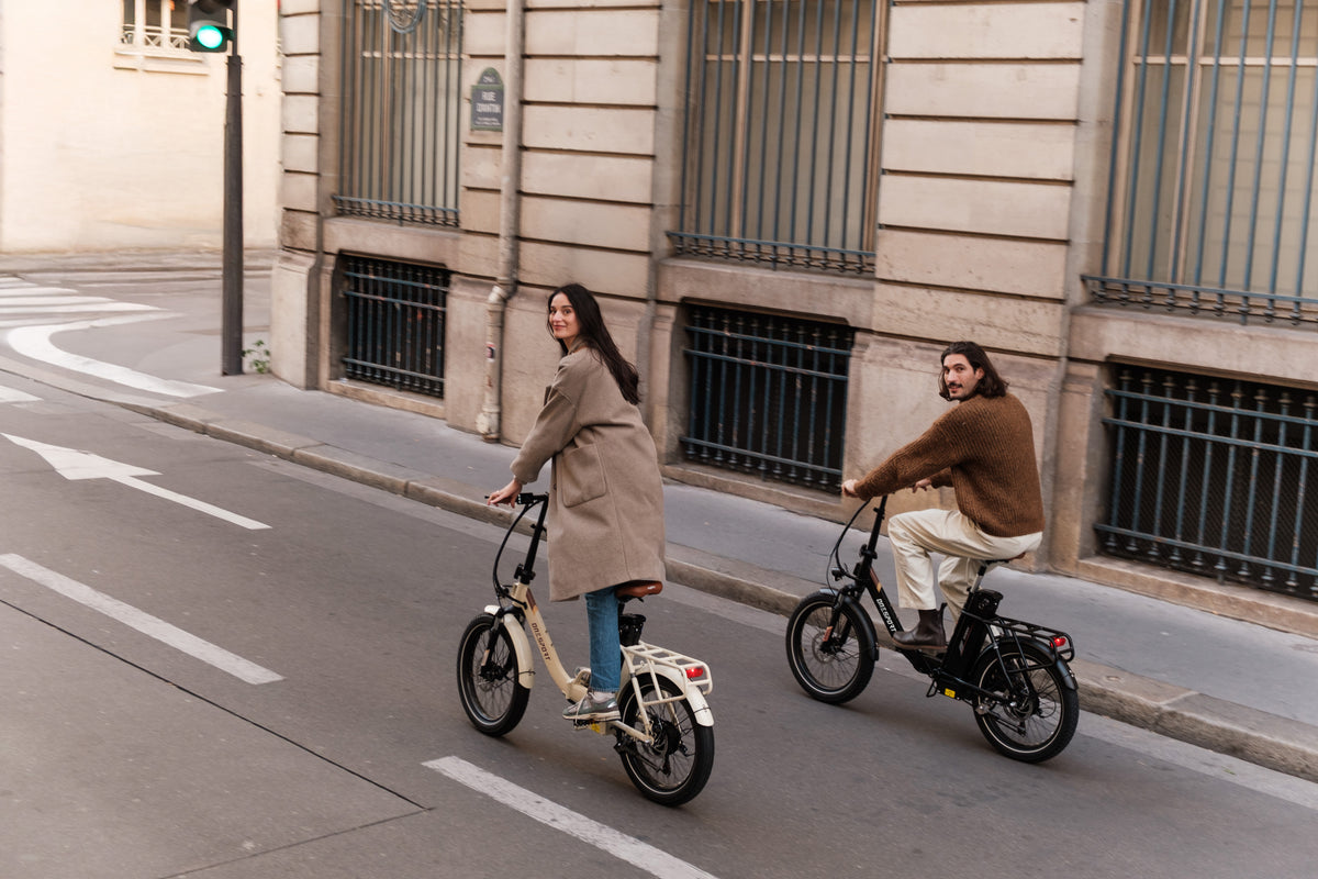 Dos personas montan en bicicleta por una calle de ciudad, cada una en una bicicleta plegable, una beige y una negra
