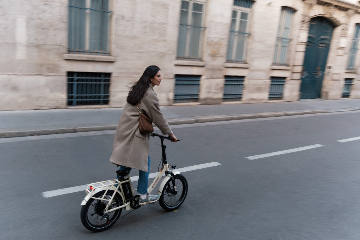 OneSport OT16 Pro: mujer con abrigo montando en bicicleta por una calle delante de una fachada de piedra, vista lateral