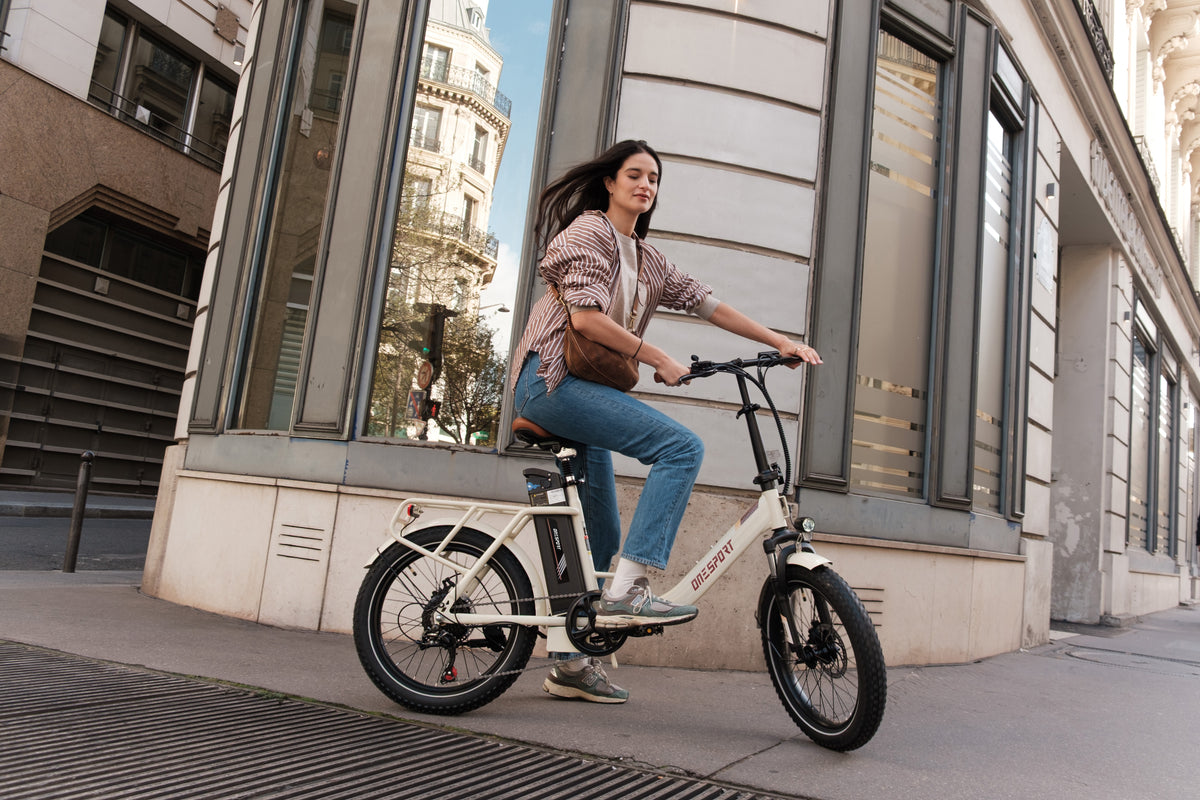 Mujer montando en bicicleta la OneSport OT16 Pro, bicicleta eléctrica blanca, vista lateral en una calle de ciudad junto a un edificio de piedra