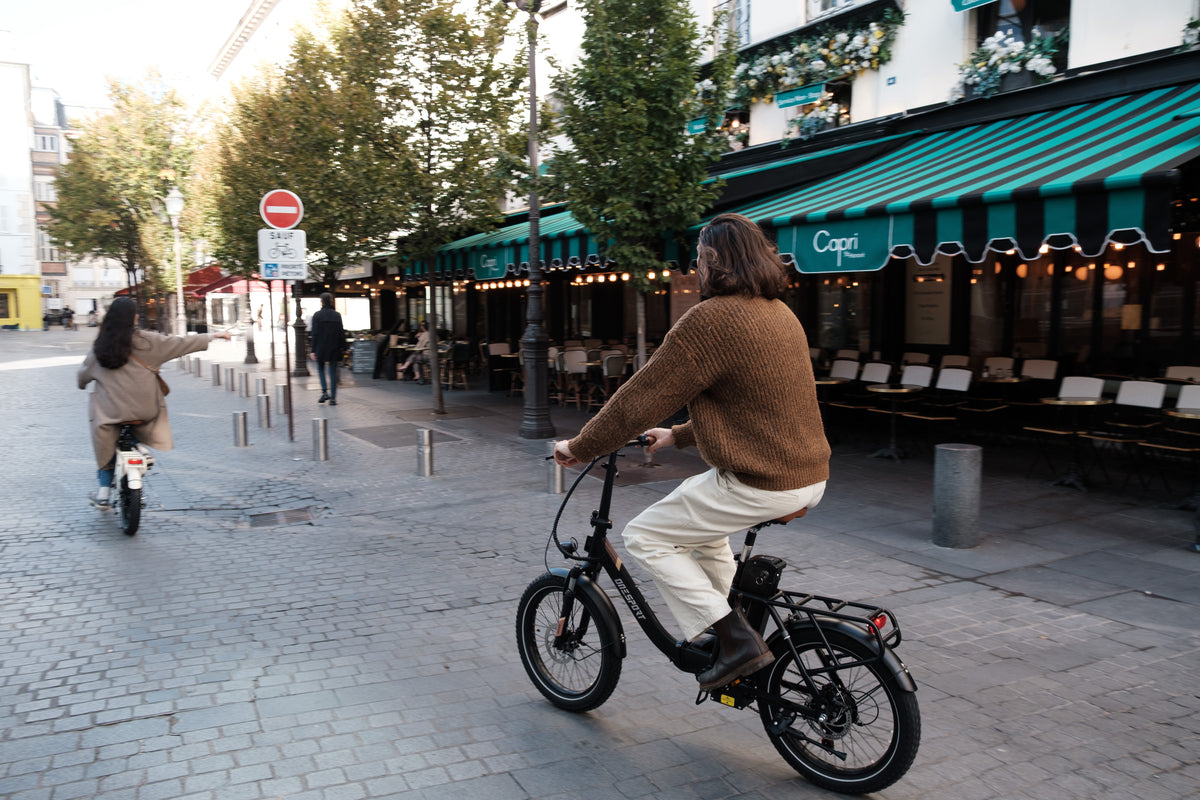 Persona montando en bicicleta la OneSport OT16 Pro, bicicleta negra, por una calle empedrada junto a un café con toldo de rayas verdes