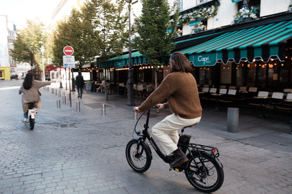 Persona montando en bicicleta la OneSport OT16 Pro, bicicleta negra, por una calle empedrada junto a un café con toldo de rayas verdes