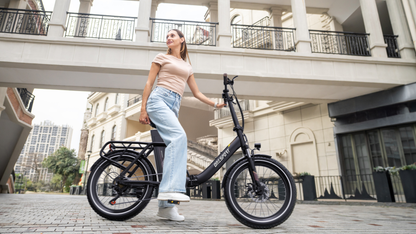 OneSport OT16 Pro: bicicleta negra en vista lateral con una mujer de pie junto a ella en una plaza adoquinada