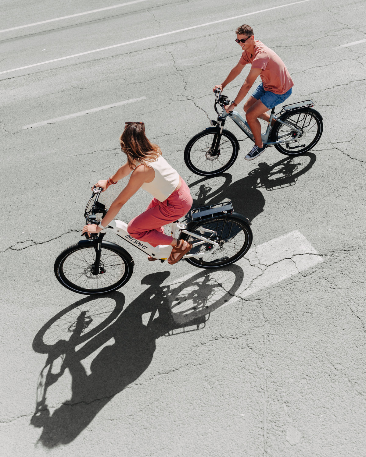 OneSport WT05 vista desde arriba, mujer en bicicleta blanca y hombre en bicicleta gris circulan sobre asfalto con sombras