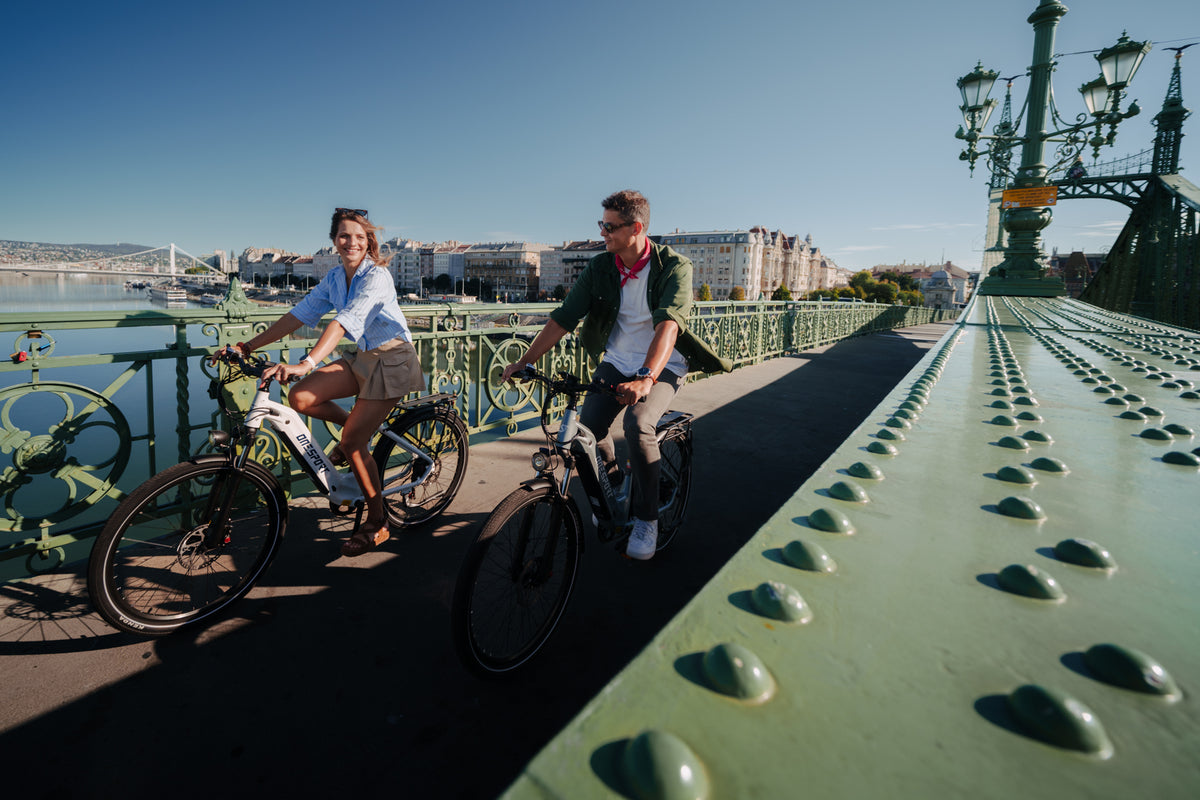 Dos personas en bicicleta sobre un puente verde bajo el sol, con edificios y agua al fondo