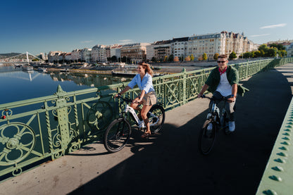 Dos personas pedalean sus OneSport WT05 sobre un puente verde con río y ciudad al fondo