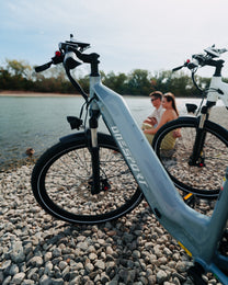 OneSport WT05 en una playa de piedras junto al agua con el cuadro en primer plano y dos personas sentadas al fondo