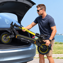 Hombre con gafas de sol levantando un patinete eléctrico negro y amarillo plegado con el texto FREEBOY junto al maletero abierto de un coche gris