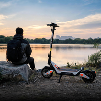 Wheely - J18 apoyado en el caballete junto a un lago al atardecer, con una persona con mochila sentada en una piedra al lado