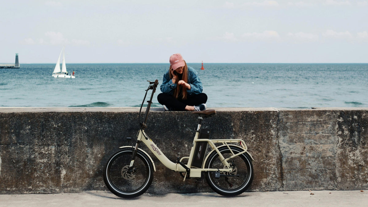 Hombre de pie junto a la bicicleta eléctrica plegable OneSport OT16-2 en color beige junto al mar