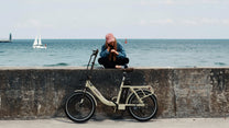 Hombre de pie junto a la bicicleta eléctrica plegable OneSport OT16-2 en color beige junto al mar