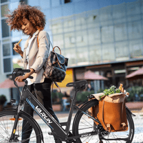 Mujer de pie junto a la bicicleta eléctrica VARUN S700-2 en color negro con cesta trasera en la ciudad